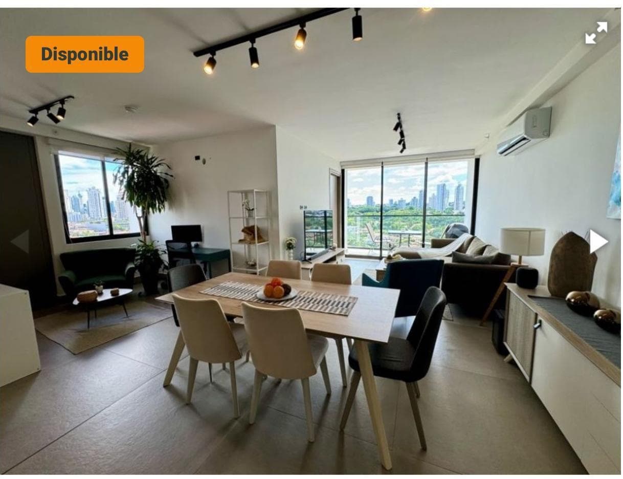 Open-plan living and dining area with table and chairs, sofa and floor-to-ceiling windows opening to a balcony with city and green views.